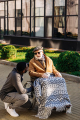happy senior disabled man in wheelchair and african american nurse chatting on street