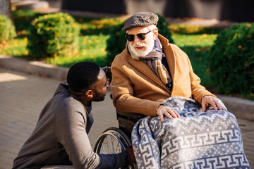 senior disabled man in wheelchair and african american nurse chatting on street