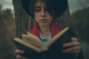 Young woman with book in her hands sits next to tree trunk and reads in dusk.
