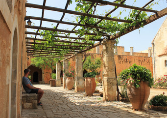 Greek courtyard - A man sits in the shade of a canopy - The yard is decorated with flowers in large pots and grapes
