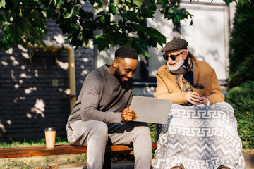 senior disabled man in wheelchair and african american man using laptop together on street