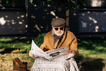 handsome senior disabled man in wheelchair reading business newspaper on street
