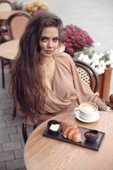 Beautiful young woman with long hair in the cafe sits at a table, drinks coffee and eat croissant. Outdoor portrait of pretty female in biege blouse enjoying latte coffee cup.