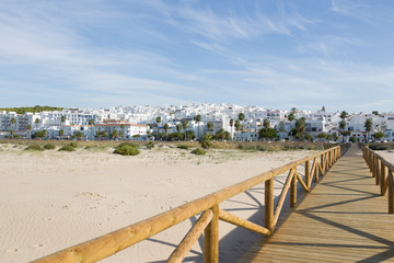Wooden boardwalk leads between palm trees to the white city of Conil de la Frontera in Andalusia with blue sky