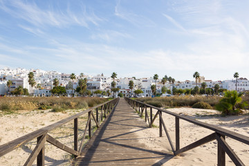 Wooden boardwalk leads between palm trees to the white city of Conil de la Frontera in Andalusia...