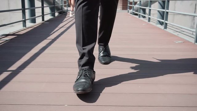 Male legs in black shoes and trousers walking over bridge. Female legs in white shoes in background. Confident walk concept