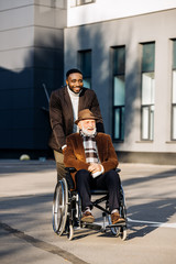 smiling senior disabled man in wheelchair and african american cuidador riding by street