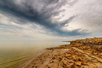 Fototapeta na wymiar The pre-storm cloudy sky above the calm sea water surface and stony shore with the groyne. Taganrog bay, Azov sea, Russia