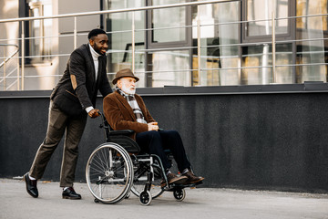 smiling senior disabled man in wheelchair and african american man riding by street