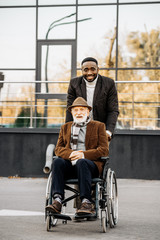 senior disabled man in wheelchair and african american man riding by street