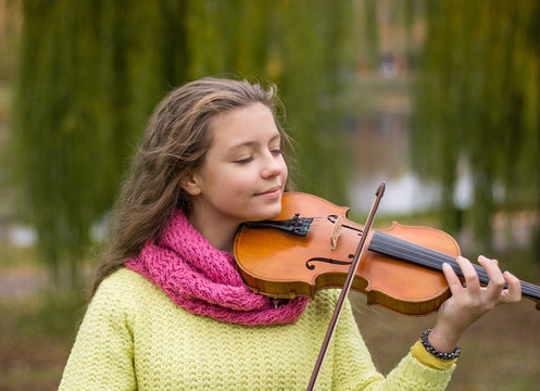 Girl playing the violin eyes closed in the autumn park at a lake and willow foliage background.