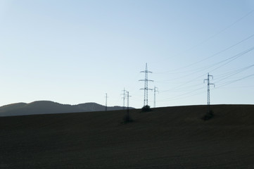Electricity pylon - pole for transmission of electricity and electric energy. Minimalist nature of field and hills around facility. Dark evening with dark ground