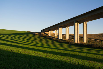 Fototapeta na wymiar Bridge, modern transport and infrastructure building. Landscape and countryside with meadow around architecture. Yellow sunset and sundown light in the evening. Shadows as stripes on the grass