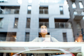 View through the window on the stylish woman sitting indoors with modern building on the...