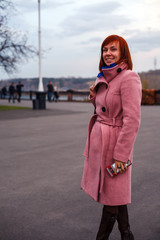 Young beautiful caucasian girl with red hair, in a pink coat with large buttons and a belt, brown boots, a colorful bag in a blue scarf. Concept of an autumn walk, a day off