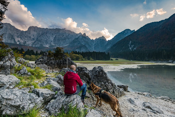 Adventure man with dog sitting at rock looking at alpine lake
