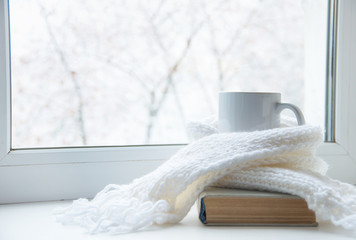 Fototapeta na wymiar Mug of hot tea and warm woolen knitting on windowsill against snow landscape from outside.