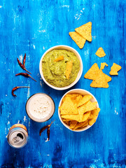 Fresh guacamole sauce in bowl with Mexican corn chips and glass of wheat beer over blue background,...