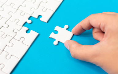 Hand putting piece of white jigsaw puzzle on blue background. Team business success partnership or...
