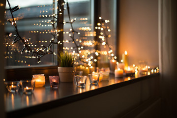hygge, decoration and christmas concept - candles burning in lanterns on window sill and festive garland string at home