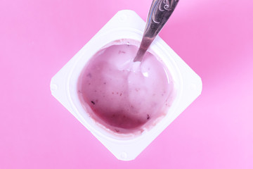 Blueberry yogurt in a plastic cup with a spoon inside on pink background closeup