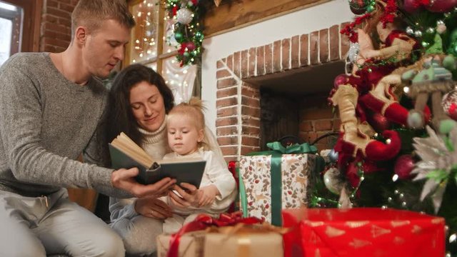 Young parents read a book to his daughter by the fireplace. The room has a lot of Christmas gifts in bright packaging and Christmas decorations. Merry Christmas 2019. Slow motion. 4K