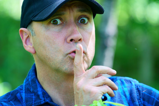 A caucasian curious man is looking away while hiking in the forest wearing a blue check shirt and a black hat.