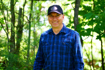Naklejka na ściany i meble A caucasian smiling man is looking into the camera for a portrait while hiking in the forest wearing a blue check shirt and a black hat.