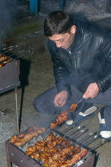 Mature man is cooking barbecued meat on mangal with skewers.