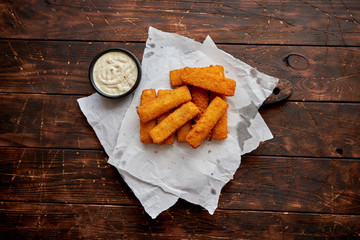 Pile of golden fried fish fingers with white garlic sauce placed on chopping board on dark wooden background.