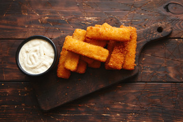 Pile of golden fried fish fingers with white garlic sauce placed on chopping board on dark wooden...