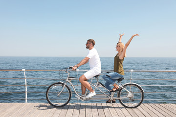 Couple riding tandem bike near sea on sunny day - obrazy, fototapety, plakaty