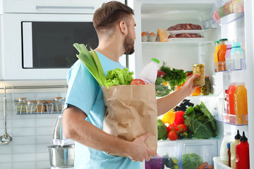 Man with paper bag putting products into refrigerator in kitchen