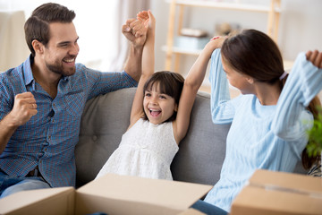 Happy multi-ethnic family with small girl sitting on couch in living room with carton boxes holding rising hands feels happy excited and glad relocate at new home. Buying moving at new house concept
