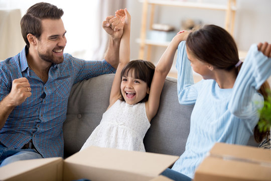 Happy Multi-ethnic Family With Small Girl Sitting On Couch In Living Room With Carton Boxes Holding Rising Hands Feels Happy Excited And Glad Relocate At New Home. Buying Moving At New House Concept