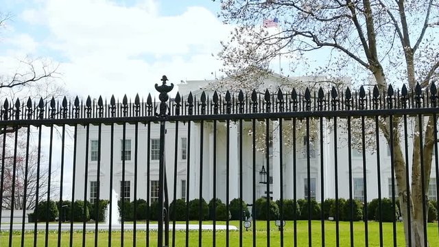 Slow motion view through fence on White House in Washington DC, USA during sunny day, blue sky, fountain splashing water in spring, green grass lawn, windy wind, American flag, nobody