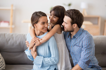 Multi-ethnic family sitting on couch in living room at home enjoy free time together on weekend communicating with each other. Beautiful preschool daughter embrace mother has fun with parents indoors