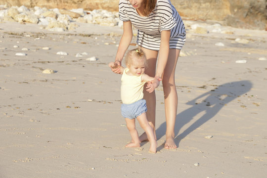 Mother helping her baby with the first steps, teaching baby to walk concept, outdoor candid photo on the beach, spending a day in the nature, healthy family lifestyle