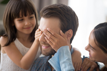 Close up happy excited caucasian man with closed eyes. Loving wife and little daughter prepare surprise for daddy husband covering his eyes with hands. Life events, father day and birthday concept
