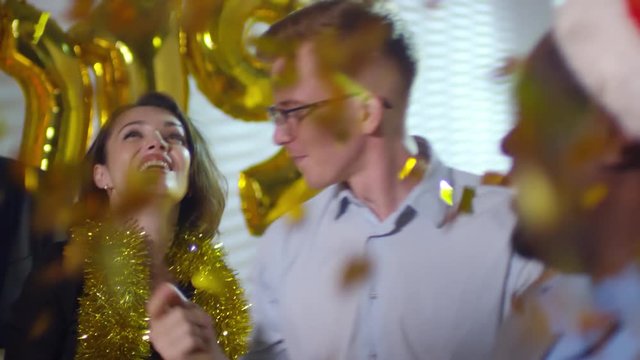 Handheld shot of happy businesswomen and businessmen dancing and cheering during New Years office party as golden confetti raining on them from above. Number 2019 balloons in background