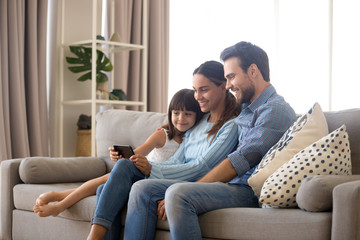 Married diverse couple spend free time on weekend with small daughter sitting on sofa in living room at home. Positive mom daddy little kid use smartphone watching cartoons videos online having fun