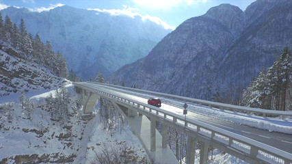 AERIAL Red car crosses the concrete bridge build in the idyllic snowy wilderness