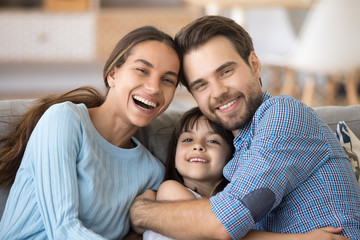 Fototapeta na wymiar Head shot portrait happy diverse cheerful laughing multi-ethnic family with little adorable sweet daughter embracing sitting on couch in living room enjoying free time together at home on weekend.