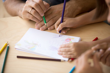 Close up people hands family sitting at table holding colored pencils drawing on paper play together at home. Kid mother and father having fun indoors. Concept of hobby and weekend leisure activities