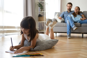 Diverse family in living room at home. Focus on little preschool adorable concentrated daughter lying at soft cushion on warm wooden floor young parents married couple sitting on sofa looking at kid