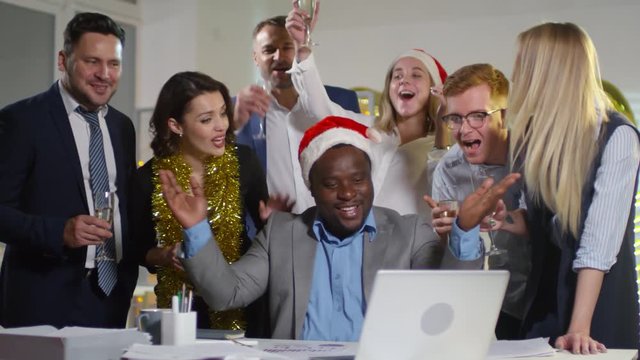 Medium shot of happy black businessman in suit and Santa hat working at his desk and closing sales deal during office Christmas party as colleagues cheering and clinking glasses of champagne