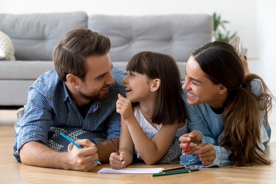 Diverse Multi-ethnic Young Family Married Couple Small Adorable Daughter Lying On Wooden Warm Floor At Modern Home Drawing On Paper Use Colored Pencils Together Have Fun Spend Free Time On Weekend.