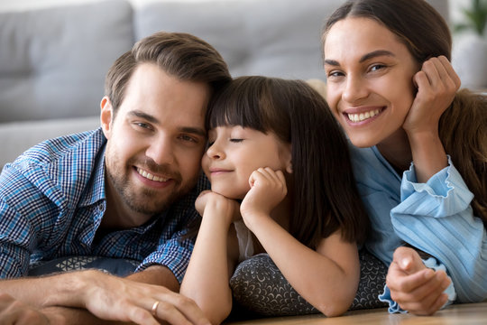 Close up little cute daughter holding hands on chin with closed eyes lying on warm floor with parents in living room at home married couple smiling and looking at camera. Happy diverse family concept