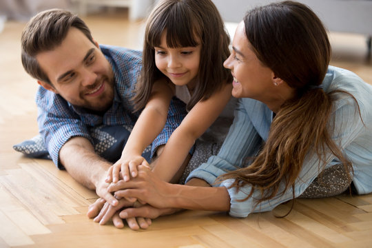 Diverse family married couple little adorable daughter lying at cushion on warm floor play smiling feels happy hold stack hands, close up. Touching arms gesture of support, love and closeness concept