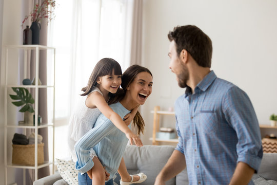 Young happy mother laughing carrying on back piggybacking little daughter catching dad playing with diverse family having fun together. Kid enjoying active games with parents in living room at home